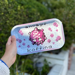 May include: A white metal rolling tray with a pink pixelated character and the text "always koffing". The tray has a pink and white checkered border.