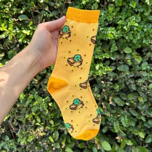 Puede incluir: Calcetines amarillos con un diseño de pato pixelado. Los calcetines tienen puños, talones y dedos amarillos. El diseño del pato es marrón y verde. Los calcetines se sostienen sobre un fondo verde y frondoso.