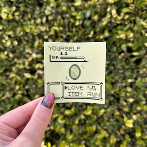 Può includere: Un adesivo verde chiaro con testo e grafica pixelata. Il testo include "YOURSELF", "L1", "HP:", "LOVE" e "ITEM RUN". Un grafico a specchio pixelato è al centro. L'adesivo è tenuto contro uno sfondo verde e foglioso.