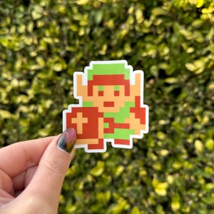 May include: A sticker featuring a pixelated video game character in green, brown, and tan. The character is holding a shield. The sticker has a white border and is set against a green, leafy background.