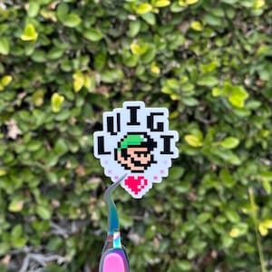 Peut inclure: Un autocollant blanc avec une image pixélisée d'un personnage au chapeau vert avec le texte "I <3  Luigi" en lettres noires.