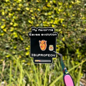 Peut inclure: Un autocollant noir avec le texte "My favorite Eevee evolution" et "IBUPROFEON". L'autocollant présente des images pixélisées d'un personnage de dessin animé et d'un flacon de médicament. Une barre de santé est en bas.