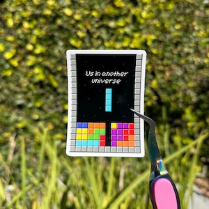 May include: A colorful sticker featuring a Tetris-inspired design with the text "Us in another universe." The sticker showcases a black background with a blue vertical line and a variety of Tetris blocks in different colors. The sticker is held by a pair of tweezers.