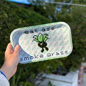 May include: A white metal rolling tray with a green and white pixelated design. The tray has the text "eat ass smoke grass" printed on it.