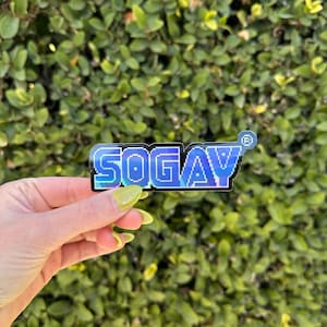 Puede incluir: Una pegatina holográfica con el texto "SOGAY" en una fuente pixelada. La pegatina es azul y negra con un efecto arcoíris iridiscente.