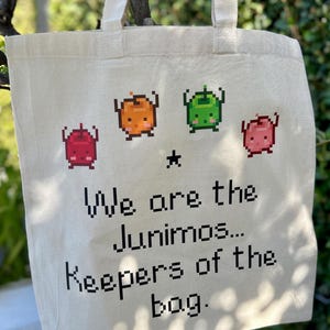 May include: A cream-coloured tote bag featuring pixelated Junimo characters in red, orange, and green. The bag has the text "We are the Junimos... Keepers of the bag." It has two handles for carrying.