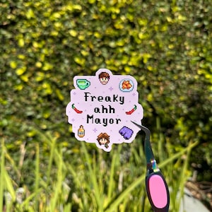 Puede incluir: Una pegatina de color lila claro con el texto "Freaky ahh Mayor" en blanco. La pegatina tiene ilustraciones pixeladas de una taza, comida, pimientos, una botella, una persona y una almohada. Unas pinzas sujetan la pegatina.