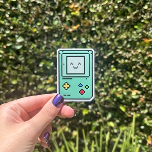 May include: A green pixelated sticker of a retro handheld video game console with a smiling face on the screen. The console has a red button, a yellow button, and two green buttons.