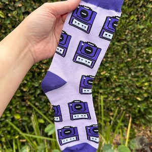 Op de afbeelding: Een paar lavendelkleurige sokken met een herhaald patroon van paarse videogameconsoles. De teen, hiel en manchet zijn effen paars. De sokken worden omhoog gehouden tegen een achtergrond van groen gebladerte.