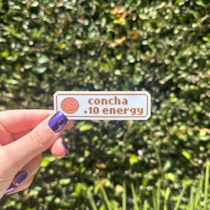 May include: A white sticker with a brown border and the text "concha +10 energy" in brown text. A pixelated image of a pink concha pastry is to the left of the text.