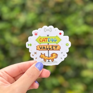 Puede incluir: Una pegatina blanca con un diseño pixelado que presenta un letrero que dice "CATDOG VALLEY". El letrero tiene una flecha verde que apunta a la izquierda y una flecha marrón que apunta a la derecha. Un gato y un perro pixelados están en la parte inferior.
