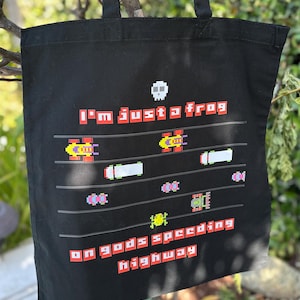 Puede incluir: Bolso tote negro con un diseño pixelado con el texto "I'm just a frog on gods speeding highway". El diseño incluye coches pixelados y una calavera. El bolso tiene dos asas negras.