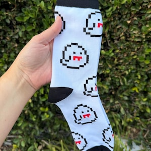May include: A white crew sock featuring a repeating pixel art pattern of ghosts. The ghosts are outlined in black with red mouths. The sock has a solid black cuff at the top, a black heel section, and a black toe section, creating a high contrast design.