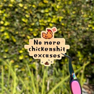 Puede incluir: Una pegatina beige con el texto "No more chickenshit excuses" y una ilustración de pollo pixelada. La pegatina está sujeta por unas pinzas con mango rosa, sobre un fondo verde borroso.