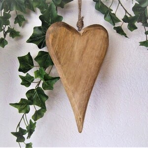 May include: A wooden heart-shaped ornament with a rustic brown finish. The heart is hanging from a twine string and is surrounded by green ivy.