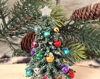 May include: A miniature artificial green Christmas tree decorated with small, colorful metallic jingle bells in red, blue, purple, and gold. The tree is topped with a small, silver glittery star and stands on a circular wooden base.