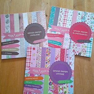 Könnte beinhalten: Drei quadratische Designpapierblöcke mit floralen und gemusterten Designs. Jeder Block ist mit einem anderen Thema versehen: Landliebe, Blumengarten und Vintage. Jeder Block ist 25 x 25 cm groß und enthält 20 Blatt beidseitig bedrucktes Papier.