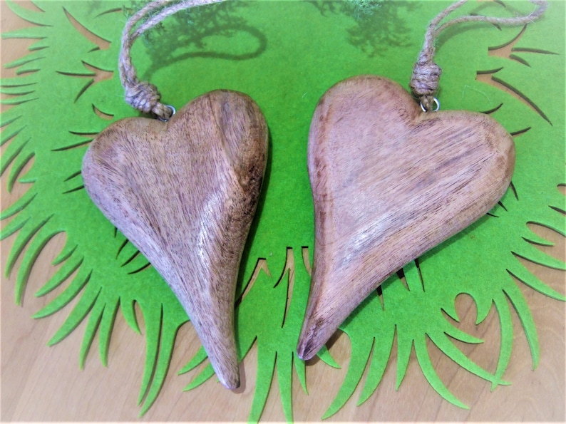 May include: Two wooden heart-shaped ornaments with a rustic brown finish, hanging from twine strings. The hearts are displayed on a green background.