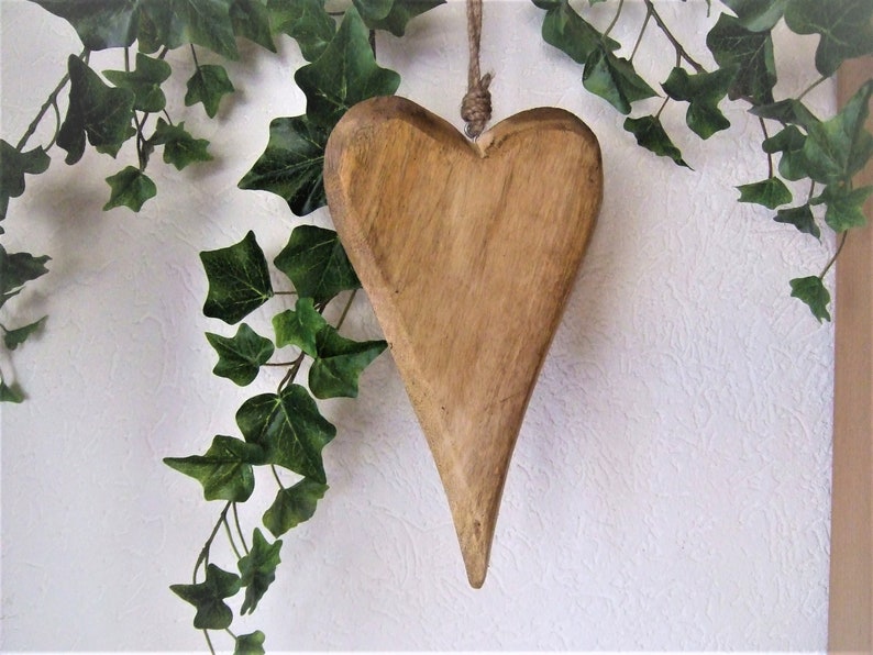 May include: A wooden heart-shaped ornament with a rustic brown finish. The heart is hanging from a twine string and is surrounded by green ivy.