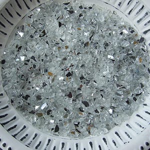 Könnte beinhalten: Eine weiße, dekorative Schale gefüllt mit kleinen, unregelmäßig geformten, klaren Glasstücken. Die Schale hat einen strukturierten Rand mit vertikalen Linien und kleinen Punkten. Die Glasstücke haben reflektierende Oberflächen.