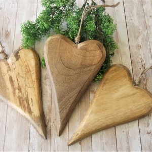 May include: Three wooden heart-shaped ornaments with a rustic brown finish. The hearts are hanging from twine and are arranged on a light wood background.