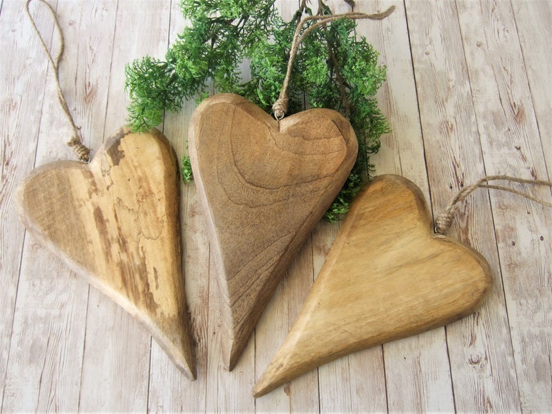 May include: Three wooden heart-shaped ornaments with a rustic brown finish. The hearts are hanging from twine and are arranged on a light wood background.