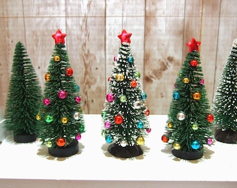 May include: Five miniature artificial Christmas trees standing in a row. Three trees are decorated with small, colorful, reflective spherical ornaments and topped with a red star. Two trees are plain green with white-tipped branches.