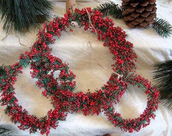 May include: Three festive Christmas wreaths with red berries and green pine needles. Each wreath is circular and has a twine loop for hanging. A pine cone and evergreen branches are in the background.