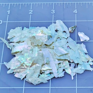 May include: A pile of iridescent white and light blue shell flakes. The flakes are various sizes and shapes. The shell flakes are on a blue grid surface.