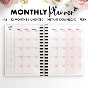 May include: A6 size monthly planner with a black and white striped spine. The planner is undated and has a pink and white color scheme. The cover features the text "MONTHLY Planner" with a heart symbol. The July page is shown with a pink heart symbol and the text "July" above the notes section. The planner has a gridded notes section and a calendar section with days of the week labeled.