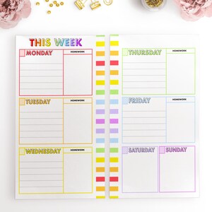 May include: A weekly planner with a rainbow color scheme. The planner has spaces for each day of the week, Monday through Sunday, with a section for homework for each day.