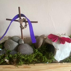 May include: A small, white, clay tomb with a pink paper butterfly on top. A wooden cross with a purple ribbon and a thorny crown is in the background. The tomb and cross are surrounded by moss and small rocks.