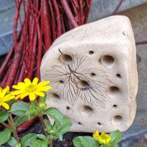 May include: A beige, stone-like sculpture with a leaf design and several holes. The sculpture is next to yellow flowers and red branches. The leaf design is etched into the surface. The sculpture has a rough, textured appearance.
