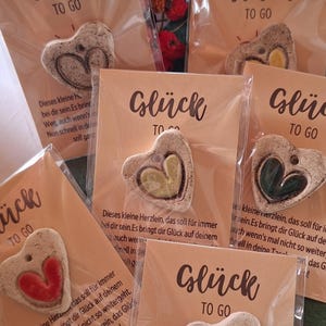 Könnte beinhalten: Fünf herzförmige Tonanhänger in durchsichtigen Kunststoffhüllen auf beigen Karten mit der Aufschrift "Glück TO GO". Jeder Anhänger hat ein zentrales Herz in Rot, Gelb, Grün oder Weiß, mit deutschem Text unter dem Anhänger auf jeder Karte.
