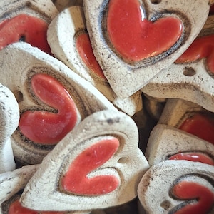 Op de afbeelding: Close-up van verschillende handgemaakte keramische hartvormige ornamenten. Elk ornament heeft een verhoogd, glanzend rood hartontwerp in een getextureerde, gebroken witte basis. De harten hebben een rustieke, ambachtelijke uitstraling.