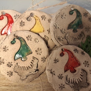 Op de afbeelding: Een verzameling handgemaakte, ronde keramische kerstversieringen. Elke versiering heeft een kleurrijk kabouterhoedontwerp in rood, geel en groen, met sneeuwvlokdetails. De versieringen zijn beige met een gat om op te hangen.