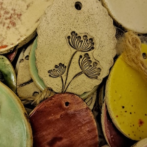 Könnte beinhalten: Eine Sammlung handgefertigter Keramikanhänger in verschiedenen Formen und Farben, darunter Gelb, Grün und Braun. Einige Anhänger weisen florale Designs auf und sind mit Naturfaser zum Aufhängen versehen.