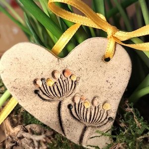 Könnte beinhalten: Ein herzförmiger Keramik-Anhänger mit rustikalem Finish, der zwei stilisierte Blumendesigns mit gelben und orangefarbenen Akzenten zeigt. Der Anhänger hat oben ein Loch zum Aufhängen und ist mit einem gelben Band gebunden.