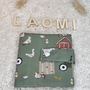 Könnte beinhalten: Ein grünes Stoffstück mit einem Bauernhoftier-Muster, darunter Hühner, Enten und eine Scheune. Das Wort "LAOMI" ist in Holzbuchstaben über dem Stoffstück geschrieben. Das Stoffstück hat eine Klappe mit einem weißen Knopf.