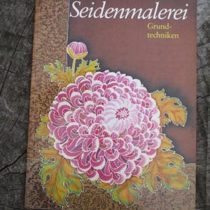 Könnte beinhalten: Ein Buch mit dem Titel "Seidenmalerei Grund-Techniken" von Ursula Kühnemnn. Der Umschlag zeigt eine rosa Chrysanthemenblüte mit grünen Blättern auf braunem Hintergrund.