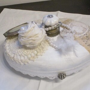 Könnte beinhalten: Weiße Keramikflasche mit einem weißen Porzellanknopf, verziert mit Spitze, Perlen und einem kleinen Spiegel. Die Flasche steht auf einer weißen Oberfläche.