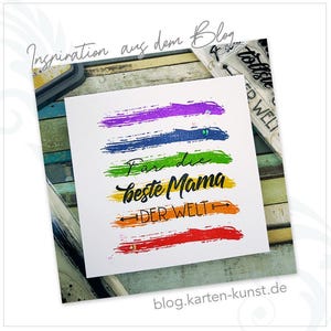 May include: A white card with rainbow colored brushstrokes and the text "F&uuml;r die beste Mama DER WELT" in black. The card is on a wooden surface.