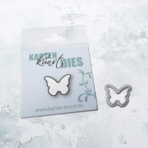 May include: A white metal butterfly die with a silver outline. The die is packaged in a clear plastic bag with a white card that says "Karten Kunst Dies" and "www.karten-kunst.de".
