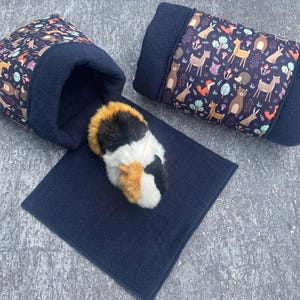 Könnte beinhalten: Zwei blaue und weiße Fleece-Tierbetten mit einem Waldtierdruck. Ein Bett ist zylindrisch geformt und das andere ist eine Tasche. Ein kleines dreifarbiges Meerschweinchen liegt im Beutelbett.