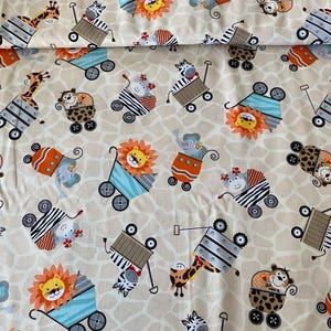 Op de afbeelding: Een beige stof met een herhalend patroon van verschillende dieren in wagens. De dieren omvatten een giraf, zebra, leeuw, olifant, aap en een beer. De wagens zijn blauw en oranje met zwarte wielen.