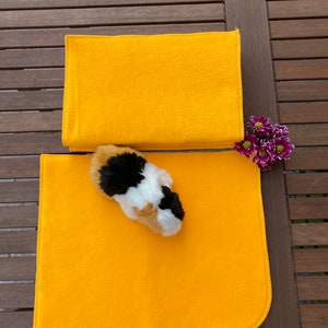Könnte beinhalten: Zwei hellgelbe Fleece-Haustierbettauflagen, eine gefaltet und eine flach, mit einem dreifarbigen Meerschweinchen-Spielzeug. Die Auflagen liegen auf einem Holztisch, mit einem kleinen Strauß lila Blumen.
