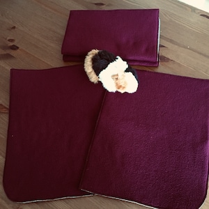 Könnte beinhalten: Ein Set aus drei bordeauxfarbenen Fleece-Artikeln, darunter zwei quadratische Matten und ein gefalteter rechteckiger Artikel, möglicherweise eine Decke oder ein Bezug. Ein kleines, flauschiges Meerschweinchen-Spielzeug mit braunem, schwarzem und weißem Fell liegt auf den Matten. Die Artikel befinden sich auf einer Holzoberfläche.