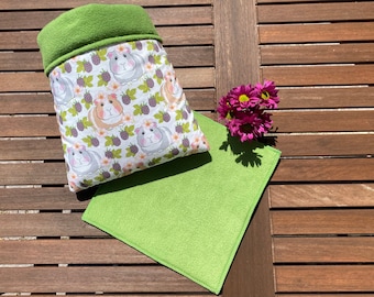 May include: Green fleece guinea pig sleeping bag with a white fabric lining featuring a pattern of guinea pigs and blackberries. The sleeping bag is on a wooden surface with a green fleece square next to it.