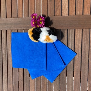 Könnte beinhalten: Drei leuchtend blaue, rechteckige Tücher sind auf einer Holzoberfläche angeordnet. Ein flauschiges, mehrfarbiges Meerschweinchen-Spielzeug liegt auf den Tüchern, mit einer kleinen Gruppe lila Blumen in der Nähe. Die Tücher scheinen aus einem weichen, saugfähigen Material zu bestehen.