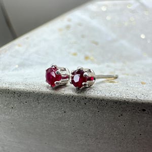 Pode incluir: Um par de brincos de pressão com pedras preciosas vermelhas vibrantes em metal prateado. As pedras são de corte redondo e presas por uma cravação de garras. Os brincos são exibidos em uma superfície cinza.